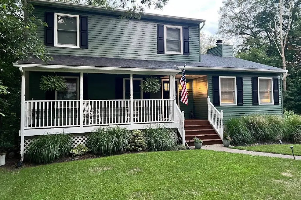 Two-story sage green colonial home featuring a white wraparound front porch with rocking chairs and an American flag.
