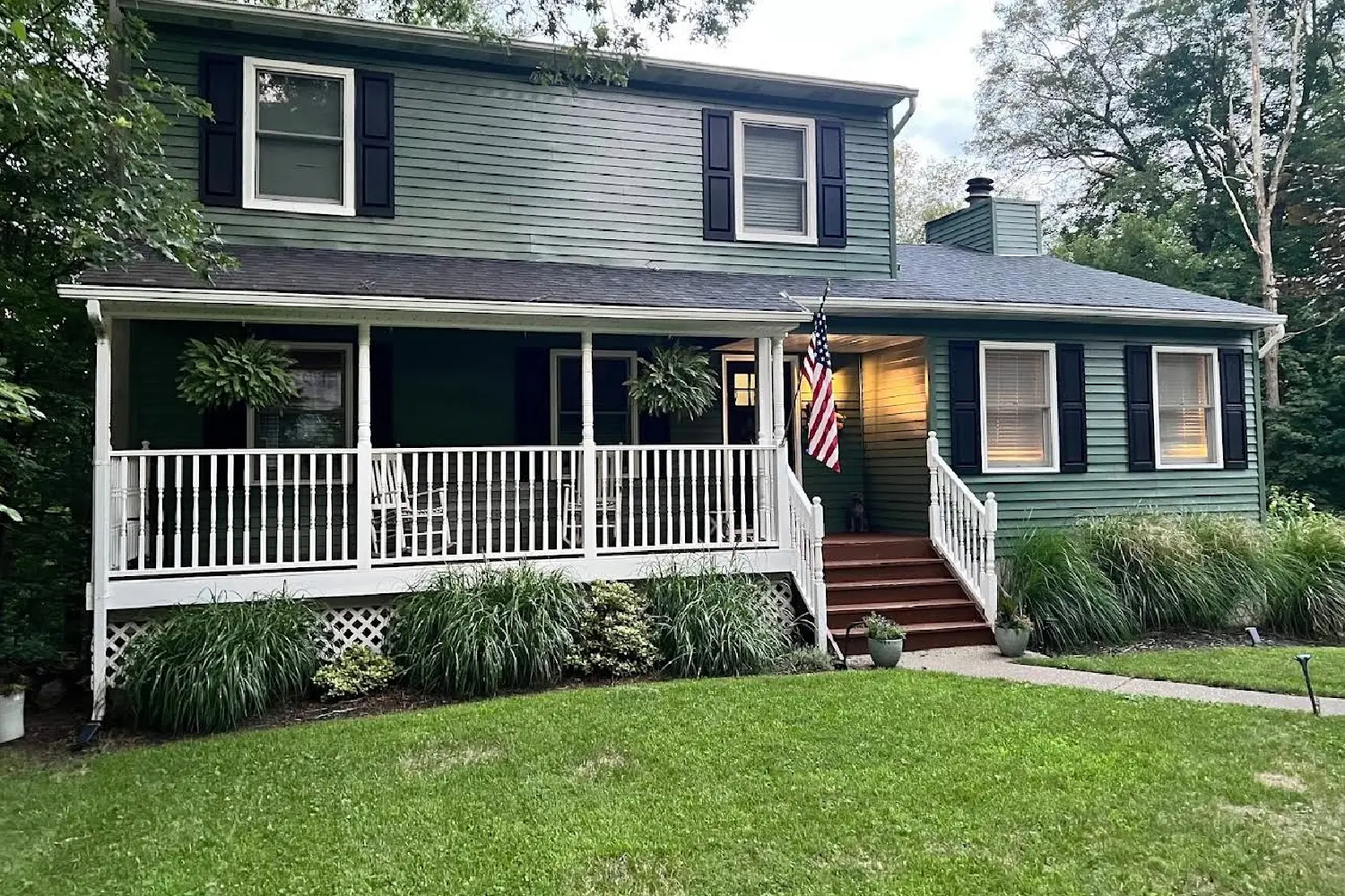Two-story sage green colonial home featuring a white wraparound front porch with rocking chairs and an American flag.
