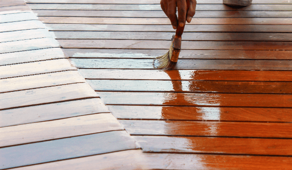 Wood deck surface being stained by hand with a brush, showing the contrast between untreated boards and freshly applied stain for long-lasting protection.
